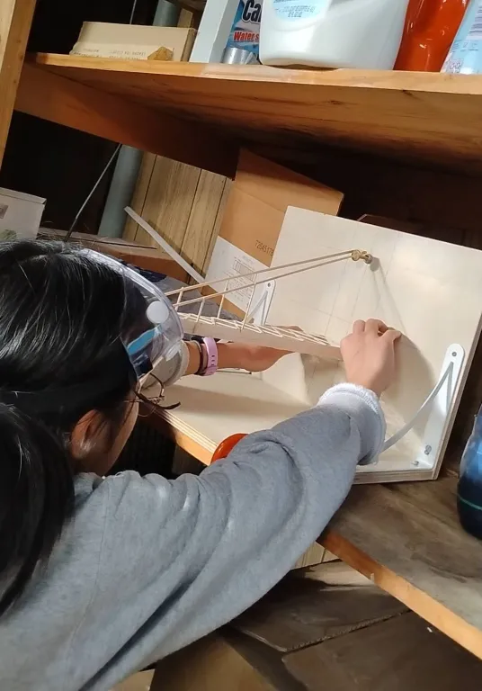 A student wearing safety goggles reaches up to adjust a wooden build-event device with a wire arm, set on a shelf.