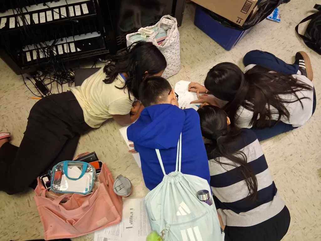 Overhead view of four middle schoolers lying and kneeling on the classroom floor in a tight circle around a worksheet, one pointing at a question.