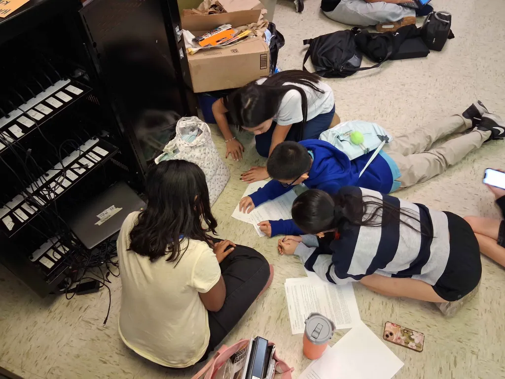 Four middle schoolers kneel and lie on the classroom floor next to a laptop cart, reading through printed test packets together.