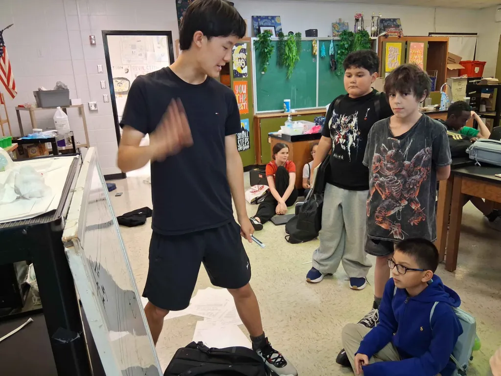 A Scioly Prep tutor in a black T-shirt points at a whiteboard while explaining a problem to three middle schoolers, two standing and one sitting cross-legged on the classroom floor.