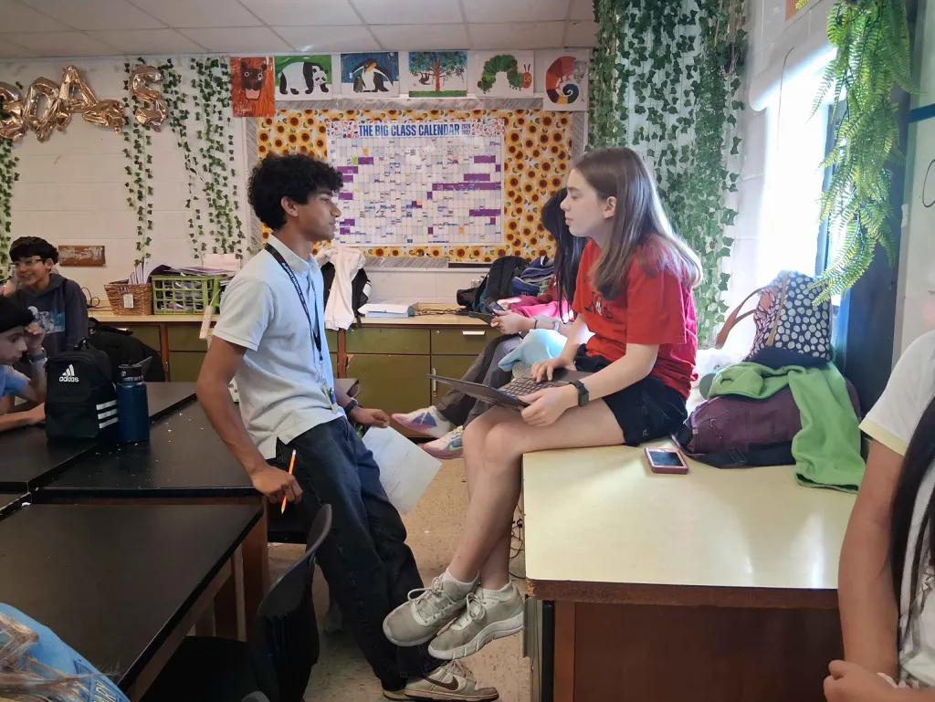 A tutor wearing a lanyard and holding a pencil talks with a student who sits on a counter with a laptop on her lap.