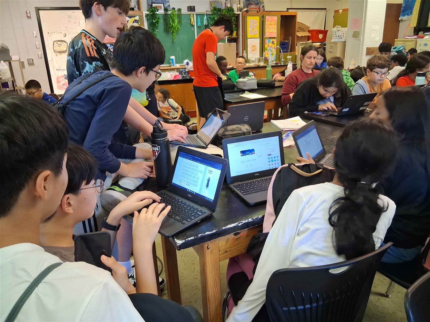 A crowded classroom during practice: middle schoolers work on Chromebooks around lab tables while an older student in an orange shirt stands among them.