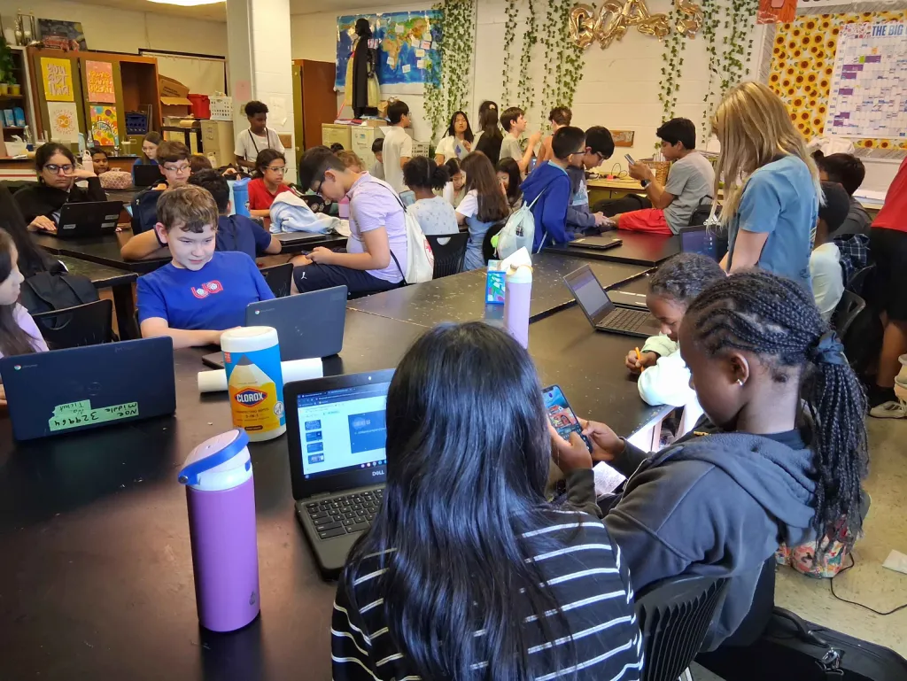 Middle schoolers work on Chromebooks around long black lab tables in a busy classroom while others stand and talk in the background.