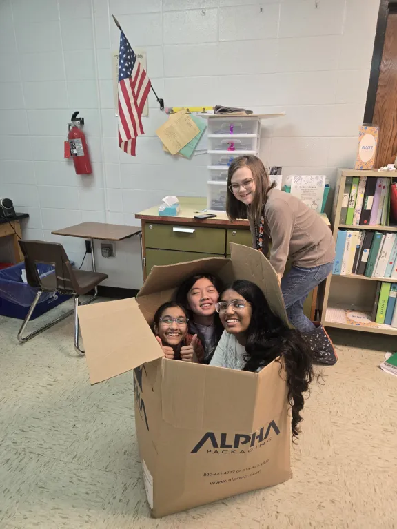 Three students squeeze into a big cardboard box in a classroom, laughing, while a fourth student leans over the side.