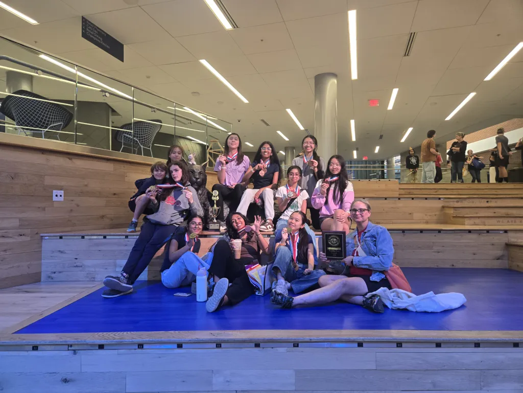 A middle school Science Olympiad team sits on blue risers at a tournament, holding up medals, a trophy, and a plaque, with an adult sitting in front beside them.