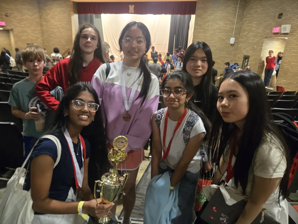 Seven middle schoolers crowd together in a school auditorium after a tournament, wearing medals on red lanyards. One kneels in front holding a gold trophy.
