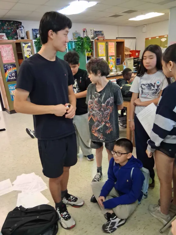 A tall tutor in a black T-shirt talks with a group of middle schoolers standing around him in a science classroom. One student in a blue hoodie sits on the floor listening.