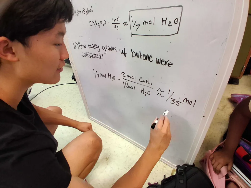 A tutor smiles while finishing a line on the whiteboard: 1/7 mol H2O times 2 mol C4H10 over 10 mol H2O is about 1/35 mol. The answer 1/7 mol H2O is boxed above it.